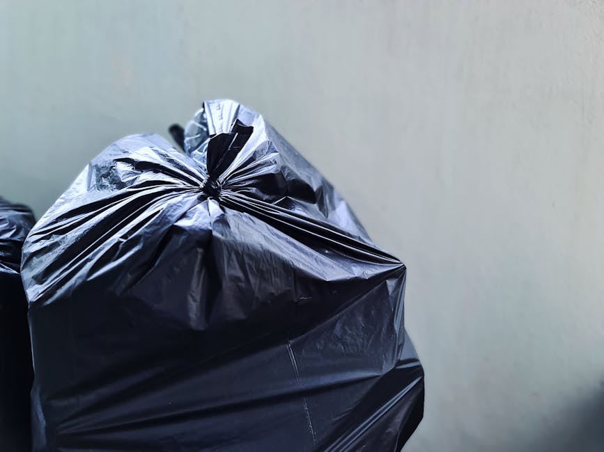 A black plastic wheelie bin situated on a street curb at night, with signs indicating it is for waste collection by St. John’s. The bin is filled with various types of rubbish, including paper, card