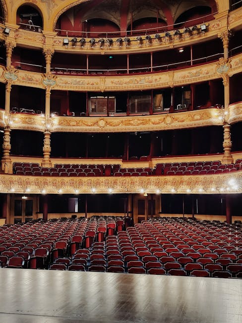 The interior of a historic theatre or opera house shows a large, ornate auditorium with multiple tiers of balconied seating, decorated with gold gilding and intricate detailing along the curved edges.